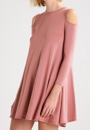 Kvinde iført en løs, støvet lyserød cold-shoulder kjole med lange ærmer, stående mod en ensartet lys baggrund.