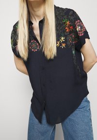 Chemise courte à manches courtes bleue marine avec broderies florales aux couleurs vives sur les épaules, boutons sur le devant et coupe décontractée.