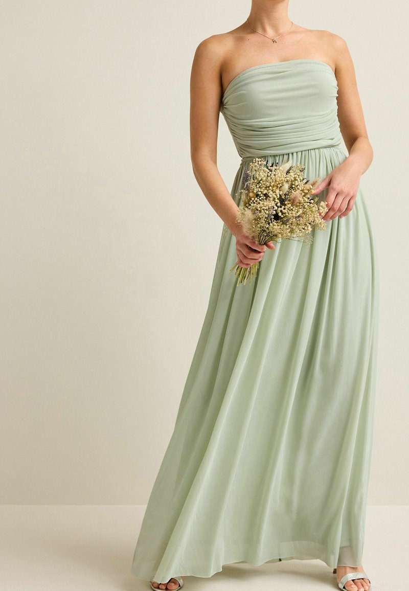 Femme en robe bustier vert clair tenant un bouquet de fleurs séchées sur un fond beige uni.