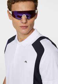 Vit polotröja med marinblå detaljer, en krage och en liten logotyp på bröstet. Ett par sportsolglasögon med lila toning och en smidig design.