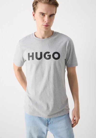 Сива памучна тениска с къси ръкави, с ярък черен текст "HUGO" на предната част. Гладка текстура и класически кръгъл деколте.