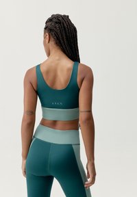 Sujetador atlético en tono azul verdoso con una banda ancha, combinado con mallas en los mismos tonos verdes y azules. La tela parece suave con un acabado mate y detalles de marca sutiles.