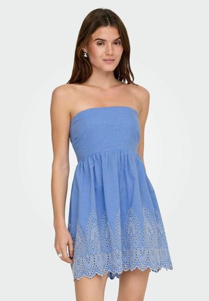 Vrouw in strapless blauwe jurk met oogjesborduursel aan de zoom, staand tegen een effen lichte achtergrond.