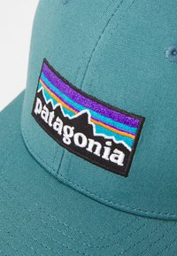 Boné teal com aba curva, apresentando uma etiqueta retangular preta. A etiqueta exibe um logotipo de montanha multicolorido e o texto "patagonia" em branco.