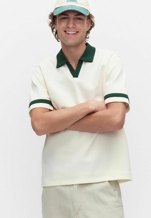 Młody mężczyzna uśmiechający się z założonymi ramionami, ubrany w kremową koszulkę polo z ciemnozielonym kołnierzykiem i mankietami, beżowe spodnie oraz jasną czapkę.