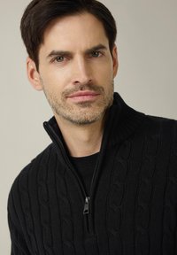 Czarny sweter z dzianiny warkoczowej z kołnierzem na zamek błyskawiczny, ozdobiony teksturowanym wzorem. Nosi się go na czarnej koszulce, podkreślając jego przytulny wygląd.