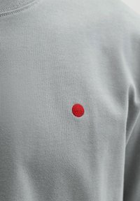 Hellgrauer Stoff mit einem kleinen, runden, roten Stickpunkt nahe der Mitte.