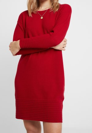 Woman wearing a red long-sleeve knit dress, arms crossed, with a gold pendant necklace against a plain light background.