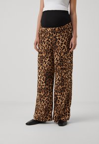 Gravid kvinna i leopardmönstrade vida gravidbyxor med svart elastisk midja och svarta spetsiga skor på neutral bakgrund.