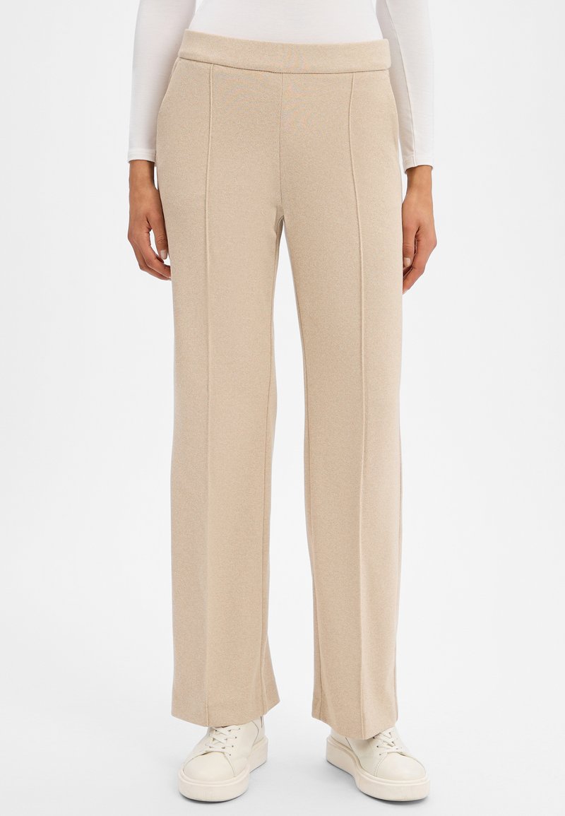 Beige Hosen mit weitem Bein, einem flachen Bund, strukturiertem Stoff und vorderen Nähten, kombiniert mit weißen Turnschuhen.