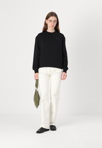 Felpa nera con polsini e orlo a coste, abbinata a pantaloni bianchi a gamba larga. Il modello tiene una borsa verde oliva e indossa scarpe nere.