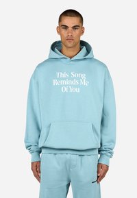 Felpa blu chiaro con testo bianco "Questa canzone mi ricorda di te." Presenta una tasca frontale e polsini a coste, abbinata a pantaloni della tuta coordinati.
