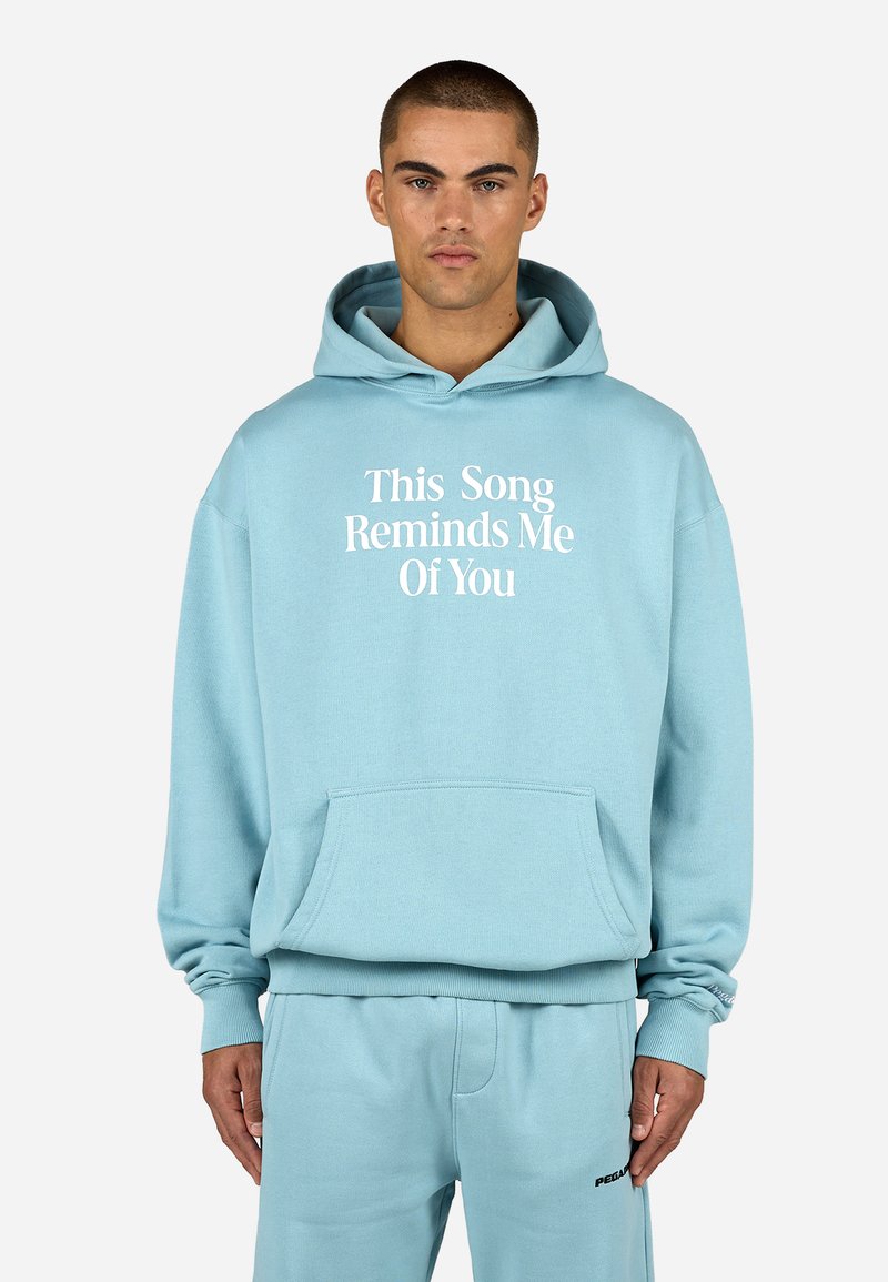 Felpa blu chiaro con testo bianco "Questa canzone mi ricorda di te." Presenta una tasca frontale e polsini a coste, abbinata a pantaloni della tuta coordinati.