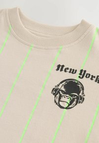 Sweatshirt beige avec des rayures verticales vert fluo et un graphique noir d'un gorille avec des écouteurs, le texte "New York" au-dessus.