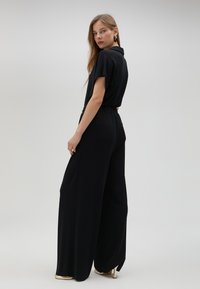 Mono negro con cuello, mangas cortas, perneras anchas y una cintura elástica para mayor comodidad. Tejido suave y ligero sin hardware visible.