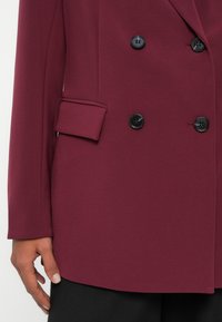 Blazer bordeaux de três quartos, com um corte estruturado, botões pretos e um único bolso lateral, feito de um tecido suave e texturizado.