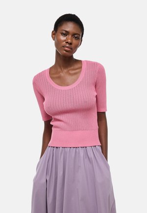 Jeune femme aux cheveux courts portant un haut rose côtelé à manches courtes et une jupe lavande taille haute, les mains dans les poches.