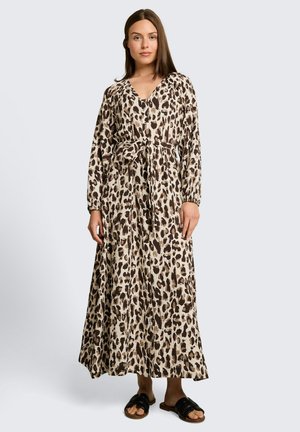 Femme portant une longue robe imprimée léopard, manches longues, ceinture à la taille et sandales noires à enfiler, debout devant un fond uni.