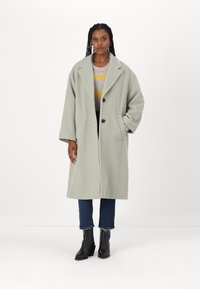 Hellgrauer oversized Mantel mit einem Reverskragen, zwei Fronttaschen und zwei schwarzen Knöpfen, getragen über einem gestreiften Pullover und dunklen Jeans.