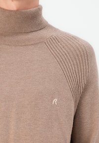 Pull à col roulé beige avec texture côtelée sur les épaules, comportant un petit logo brodé sur la poitrine. Fabriqué en tissu doux.