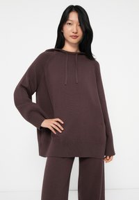 Jeune femme aux longs cheveux noirs portant un sweat à capuche en tricot oversize de couleur violet foncé et un pantalon large assorti, sur un fond blanc.