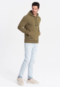 Sudadera verde oliva con bolsillo frontal, capucha con cordón y puños acanalados, combinada con jeans azul claro y zapatillas beige. Diseño casual.