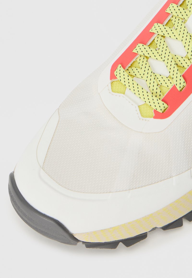 Scarpa sportiva bianca con una tomaia texture, lacci giallo neon e dettagli rossi, caratterizzata da una suola in gomma nera e un motivo a battistrada giallo a contrasto.