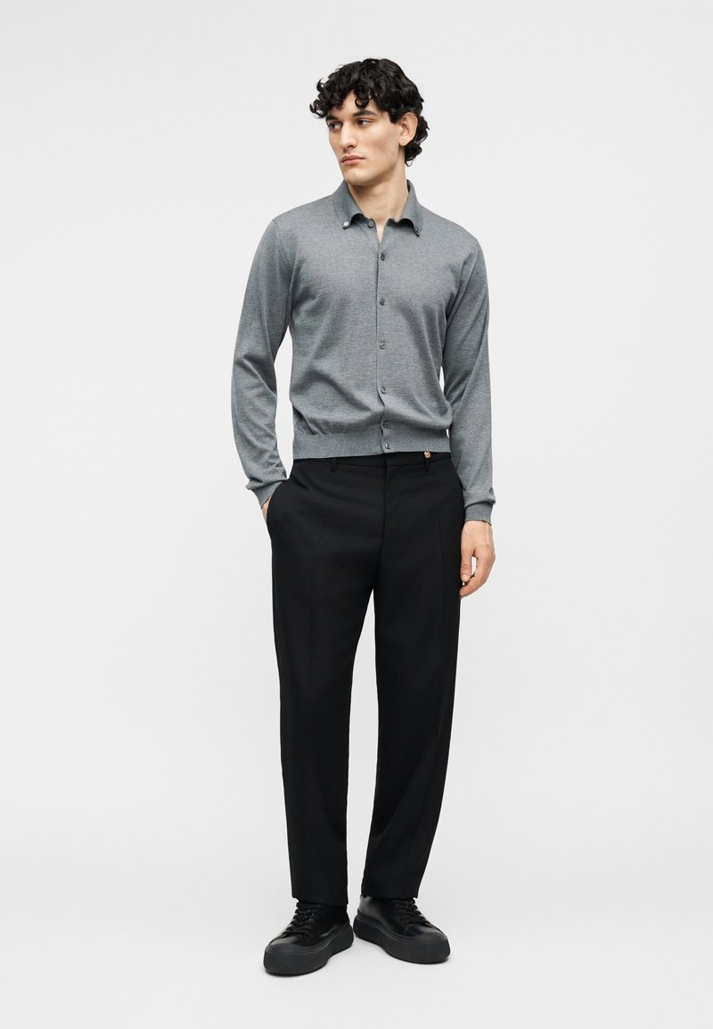 Man draagt een grijs vest met knopen, zwarte broek en zwarte sneakers, staat met één hand in de zak tegen een effen achtergrond.