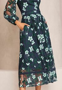 Marineblauwe geborduurde jurk met bloemenpatronen, transparante mouwen en elastische taille met zakken, die een gelaagde rokontwerp benadrukt.