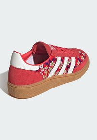 Zapatilla roja de Adidas con rayas blancas, patrón floral en la parte superior de tela, talón de ante, suela de goma y lettering dorado "Spezial" en el costado.