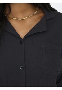 Black button-up shirt with a textured fabric, featuring a collar, single chest pocket, and a smooth, shiny button. Gold twisted necklace adorns the neck.