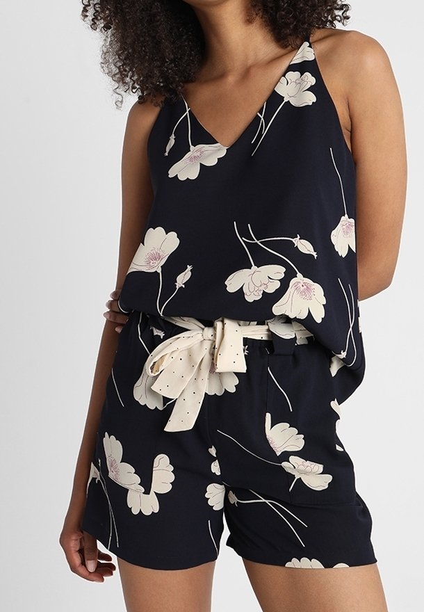 Femme portant un haut noir sans manches et un short assorti avec un imprimé floral blanc et une ceinture en tissu blanc nouée en nœud.