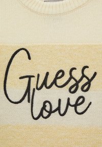 Maglione lavorato a maglia con una sfumatura tra crema e giallo pallido, decorato con un audace ricamo nero che recita "Guess love". Tessuto morbido e collo rotondo.