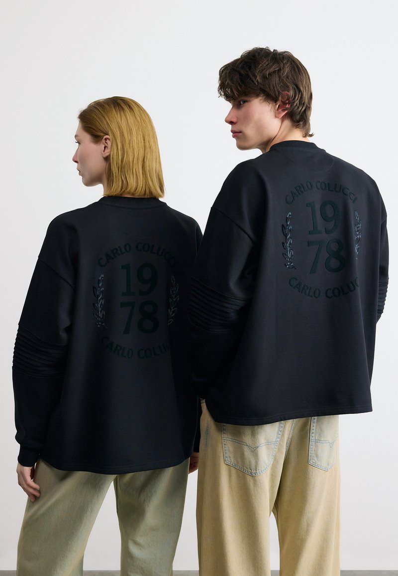 Δύο μοντέλα που φορούν μαύρα φούτερ με το κείμενο "CARLO COLUCCI 1978" και γραφικά δάφνης στην πλάτη, σε συνδυασμό με ανοιχτό μπεζ φαρδιά παντελόνια.