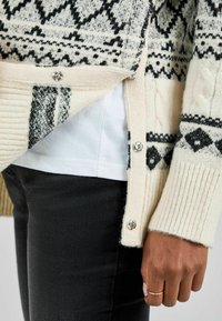 Strickjacke in Creme und Schwarz mit geometrischen Mustern, gerippten Bündchen und Druckknöpfen. Darunter ist ein weißes Hemd sichtbar.