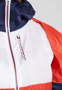 Kurtka z granatowym kapturem, białą częścią przednią i pomarańczowymi rękawami, z pionowym logo Tommy Sport na klatce i zamkiem w kolorach czerwono-biało-niebieskich.