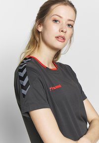 Jeune femme aux cheveux blonds portant un t-shirt de sport noir Hummel avec des bandes en chevron sur l'épaule sur un fond uni.