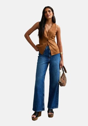 Chaleco de ante marrón, jeans de mezclilla azules holgados, sandalias de plataforma gruesa y un bolso estampado. Fondo liso, modelo en una pose relajada.