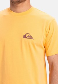 Žluté bavlněné tričko s malým purpurovým logem na levé hrudi. Klasický design s kulatým výstřihem a krátkými rukávy, hladký povrch.