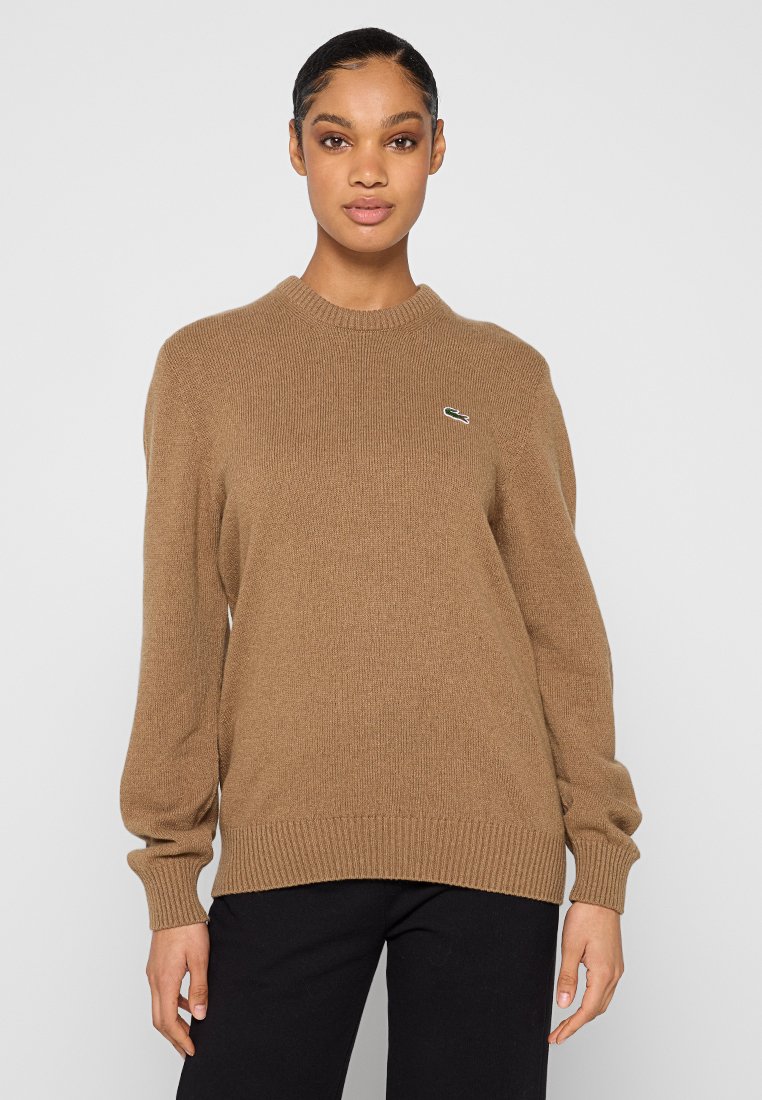 Donna che indossa un maglione lavorato a maglia color cammello con un piccolo logo di coccodrillo verde sul petto e pantaloni neri, in piedi contro uno sfondo semplice.