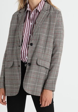 Femme portant un blazer à carreaux gris par-dessus une chemise blanche à rayures marron avec boutons, et un pantalon noir, debout devant un fond uni.