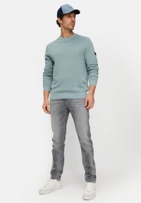 Lichtblauwe gebreide trui, ronde hals, geribbelde manchetten. Grijze spijkerbroek, witte sneakers. Donkerblauwe pet met logo. Model staand, handen ontspannen.