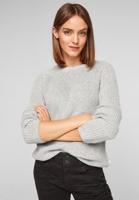 Pull en tricot gris clair avec un col rond et des manches trois-quarts, doté d'un motif texturé et d'accents côtelés, associé à un jean noir.