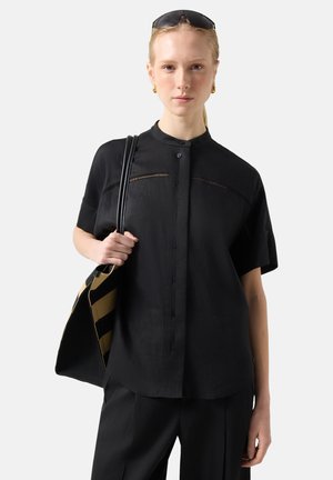 Frau trägt ein lockeres, schwarzes kurzärmeliges Hemd mit Knöpfen, schwarze Hose, Sonnenbrille auf dem Kopf, hält eine schwarz-gelb gestreifte Einkaufstasche.