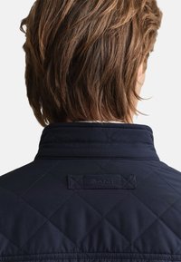 Temno modra prešita jakna z visokim ovratnikom, s teksturiranim vzorcem in znamkasto značko na hrbtni strani. Oblikovana iz gladkega materiala.