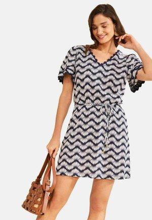 Žena oblečená v krátkych tmavomodro-bielych šatách so vzorom mušlí a zvlnenými lemami, drží hnedú pletenú koženú kabelku.