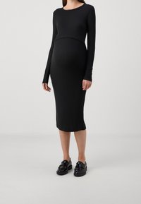 Svart randig gravidklänning med lång ärm och rundad halsringning. Fitted silhuett som slutar strax under knäet, tillsammans med svarta loafers.