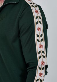 Zelená bunda na zips s bielymi bočnými pruhmi, na ktorých je vyšitá červená ruža a zelené listy. Mäkký materiál, priliehavý strih.