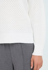 White knitted sweater featuring a textured open weave pattern and ribbed cuffs, paired with gray fabric pants, showcasing a relaxed fit.