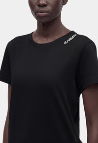 Femme portant un t-shirt noir uni à manches courtes avec un petit logo blanc « armani » brodé près de l'encolure, côté gauche.